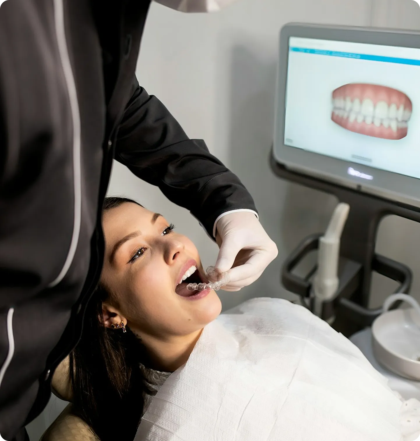 Dentist fitting a clear aligner onto a smiling patient's teeth, with a digital scan displayed on a monitor behind them